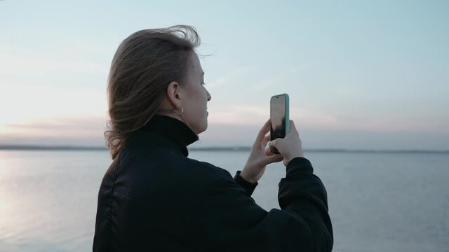 A woman takes pictures on her phone of a sunset on a lake on an autumn evening. Taking video of a sunset over the lake. Woman loves nature. High quality 4k footage