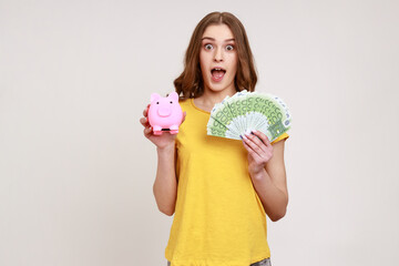 Extremely shocked female of young age in yellow T-shirt holding piggy bank and euro banknotes...
