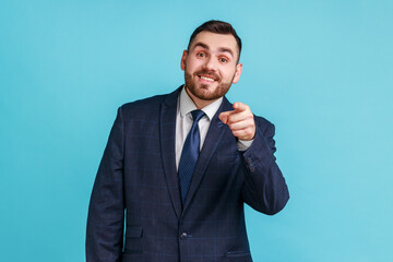 You are special! Young adult man with beard wearing official style suit pointing fingers at camera and looking with toothy smile, believes in you. Indoor studio shot isolated on blue background.