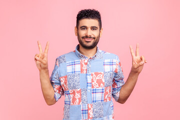 Fototapeta premium Happy successful man with dark hair and beard in blue casual shirt showing v sign victory gesture with fingers, looking at camera with smile. Indoor studio shot isolated on pink background.
