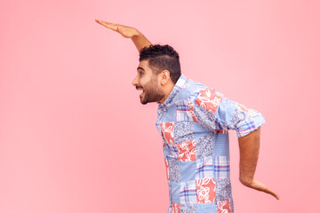 Side view of cheerful bearded man in blue casual shirt dancing, having fun and doing funny egyptian arm dance move, celebrating, rejoicing. Indoor studio shot isolated on pink background.