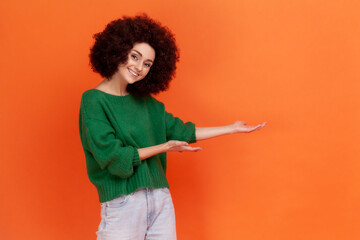 Happy friendly woman with Afro hairstyle wearing green casual style sweater presenting advertising area with toothy smile, copy space. Indoor studio shot isolated on orange background.