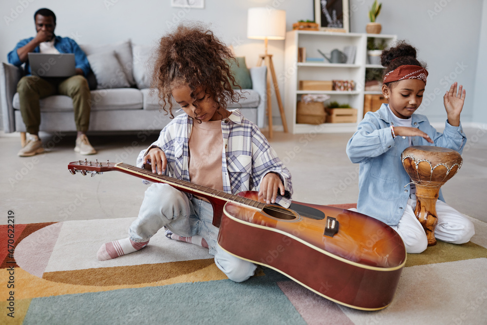 Wall mural Full length portrait of two African-American girls playing with musical instruments on floor in cozy home interior