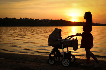 Obraz na płótnie Canvas Happy mother with baby in stroller walking near river at sunset