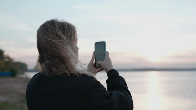 A woman takes pictures on her phone of a sunset on a lake on an autumn evening. Taking video of a sunset over the lake. Woman loves nature. High quality 4k footage