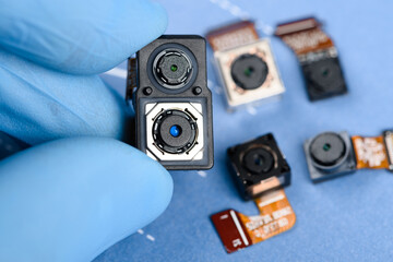 Smartphone dual sensor camera module in scientist hands, with other cell phone cameras on background.