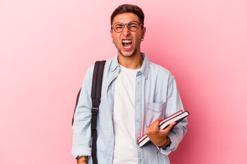 Young caucasian student man holding books isolated on pink background  screaming very angry and...