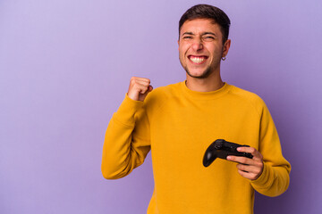 Young caucasian man holding game controller isolated on purple background  raising fist after a victory, winner concept.