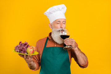 Portrait of bearded handsome dreamy grey-haired man chef drinking wine eating snack isolated on vivid yellow color background