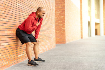 Sportsman wearing a red jacket leaning against a brick wall of clutching his chest acute pain possible heart attack. Hands male holding chest with symptom heart attack disease on city street.