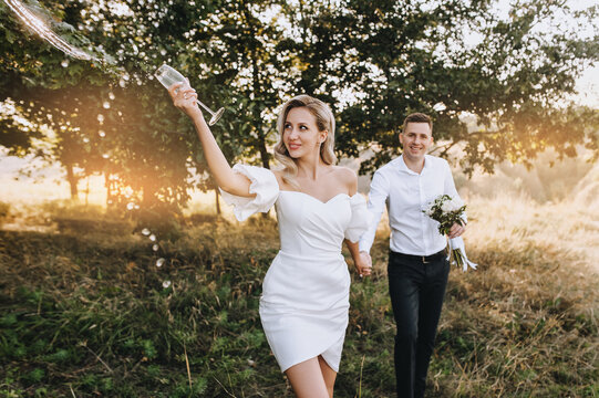 A young, beautiful blonde bride pours out champagne from a glass while walking by the hand with the groom in nature at sunset. Wedding photo of cheerful newlyweds, idea.