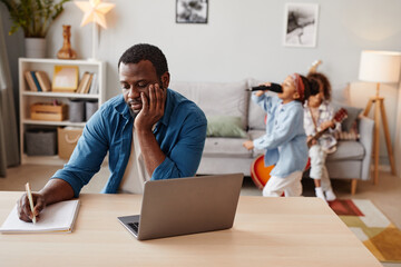 Portrait of African-American man using laptop while working at home with kids playing in background, copy space