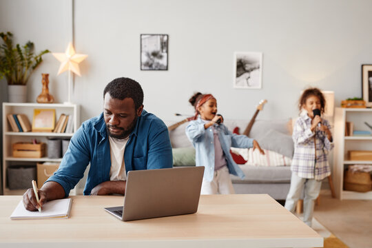 Portrait of African-American man using laptop while working at home with children playing in background, copy space