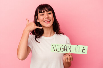 Young caucasian woman holding a vegan life placard isolated on pink background showing a mobile...