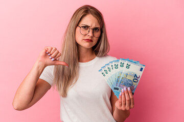 Young russian woman holding bills isolated on pink background showing a dislike gesture, thumbs...