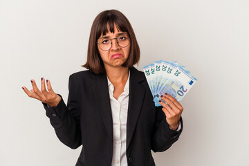 Young mixed race business woman holding a banknotes isolated on white background shrugs shoulders and open eyes confused.