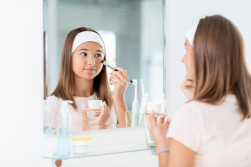 Obraz na płótnie Canvas beauty, make up and cosmetics concept - teenage girl with brush applying face mask and looking to mirror at home bathroom