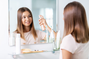 Obraz na płótnie Canvas beauty and people concept - teenage girl looking to mirror and using hair styling spray at home bathroom