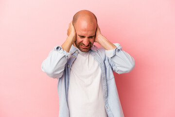 Young caucasian bald man isolated on pink background  covering ears with hands.