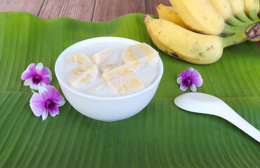 Traditional Thai dessert, bananas in coconut milk, in white bowl on banana leaf background.