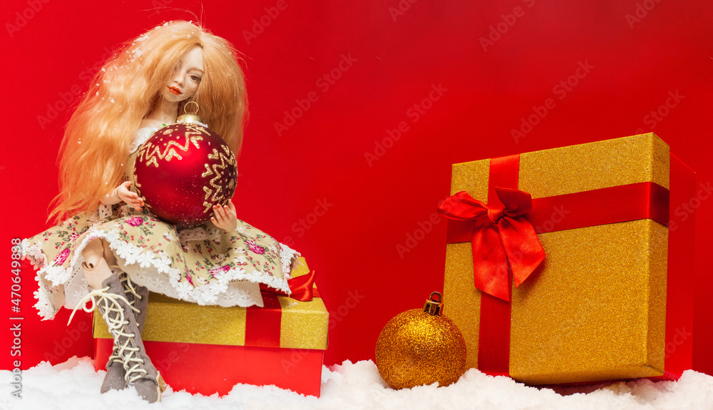 Wall mural doll with a Christmas ball in her hands sits on a New Year's gift on a red background