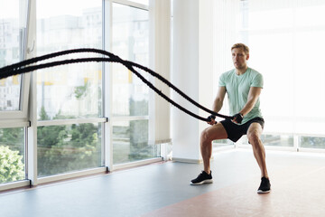 Fototapeta na wymiar Caucasian blonde man training with rope in functional training fitness gym with big windows and a lot of daylight.