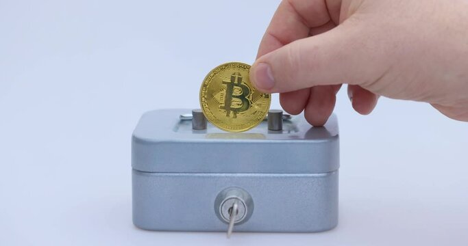 Male hand puts a cryptocoin, Bitcoin or BTC, in a small grey vault, safe or piggy bank, depicting crypto or cryptocurrency saving, holding or hodling. High quality 4k footage