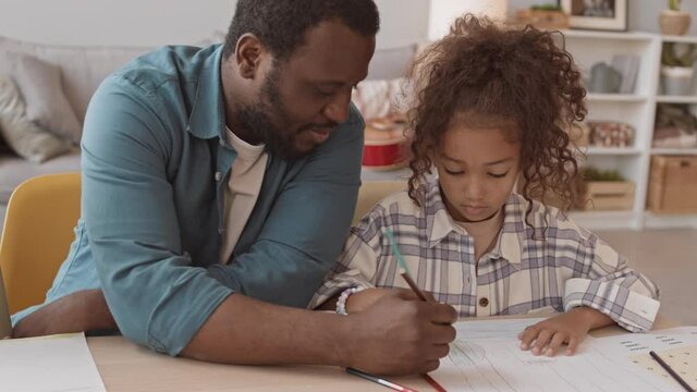 Tracking waist-up slowmo shot of African-American man sitting at table between two little daughters helping them with drawing, spending leisure time together at home
