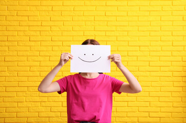 Woman hiding behind sheet of paper with happy face against yellow brick wall