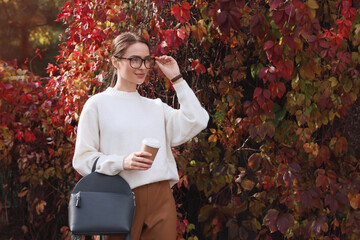 Beautiful young woman with stylish grey backpack and hot drink in autumn park