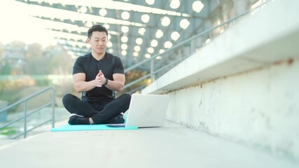 handsome asian fitness coach holds workout classes online using video call webcam and laptop. sitting outside in a modern stadium. Pilates training or stretching. Physical exercises. Records Lessons