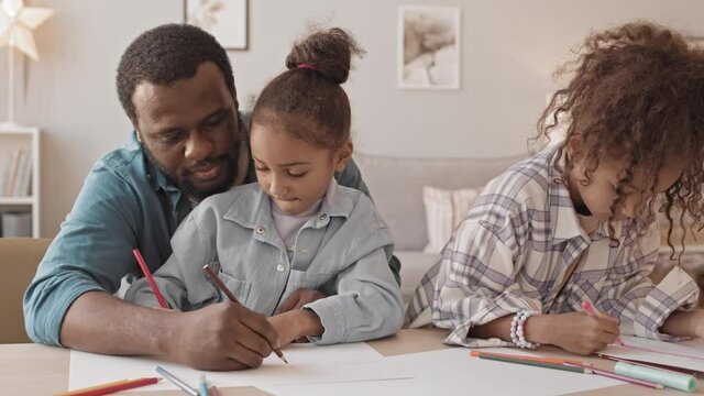 Medium slowmo shot of cheerful African-American man and his two lovely daughters drawing together sitting at table in cozy living room