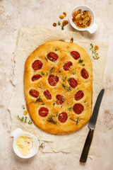 Italian traditional focaccia bread baking with with cherry tomatoes, parmesan and rosemary on light brown background. Top view