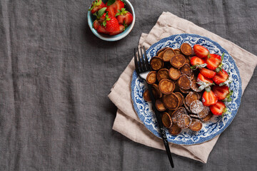 Mini chocolate pancake cereal with strawberries for breakfast on gray old textile tablecloth. Trendy home breakfast with tiny pancakes. Top view