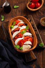 Caprese salad Italian food with ripe tomatoes, fresh garden basil and mozzarella cheese in olive wooden bowl on old rustic background. Top view flat lay