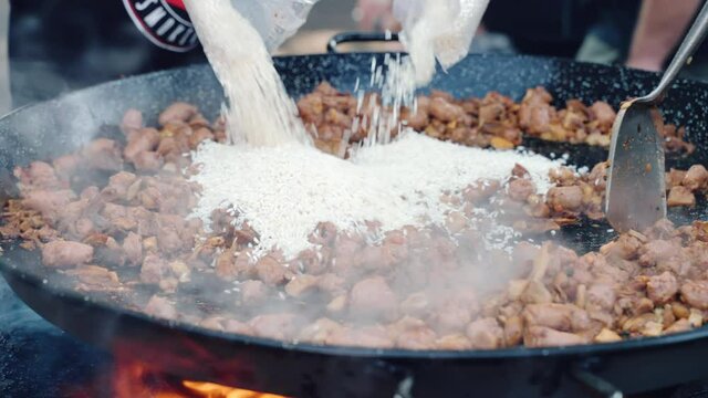Big metal cauldron on burning fire, rice pouring intro hot cauldron with frying sausages and chicken pieces. Street food preparation, cooking dishes in cauldron