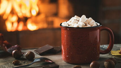 Cocoa with marshmallows and chocolate in a red mug on a wooden table near a burning fireplace, horizontal banner