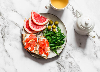 Delicious breakfast - salmon cream cheese, grain bread sandwiches, arugula, olives, grapefruit slices and green tea on a light background, top view