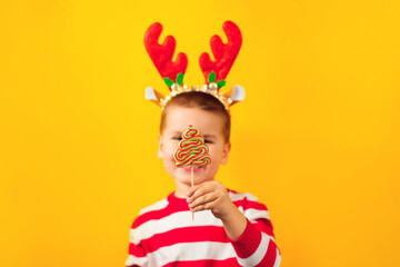 Funny child in a Santa hat with deer antlers with candy tree , yellow background. Holidays, Traditions, Christmas concept.