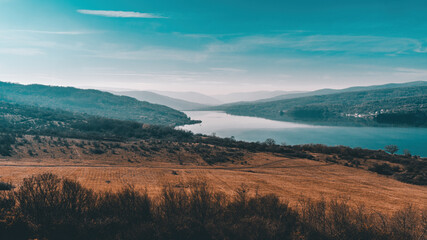 Landscape of a lake next to bare yellow field and mountains in the distant background. Sunny autumn day with blue sky