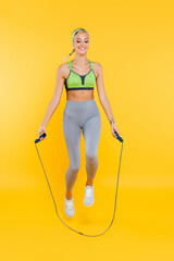 full length view of happy woman in grey leggings and green sports bra jumping with skipping rope on yellow