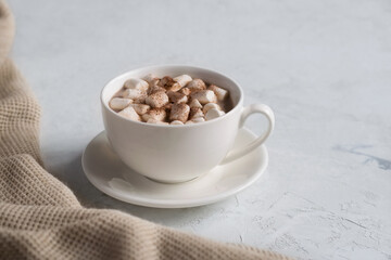 Hot chocolate with marshmallows in a white cup surrounded by a warm blanket on a light table. The concept of a cozy rest. Copy space. Selective focus.