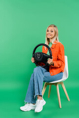 full length view of smiling blonde woman in trendy clothes sitting on chair with steering wheel on...