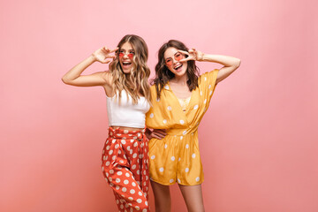 Photo of two young caucasian women in bright clothes with polka dots standing on pink background....