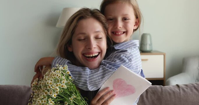 Portrait happy joyful millennial mother cuddle little daughter hold bouquet of daisies card with drawn heart. Excited mom hug small girl look at camera thankful for present on spring holiday Women Day