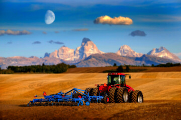 Fototapeta premium Tractor Farming Ground Harvesting Crops in Fall Autumn Teton Mountains Rugged with Moon Blurred