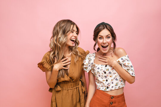 Happy young fair-skinned girls laugh while enthusiastically holding their breasts with their hand. Standing on pink background, teenagers with curly hair are dressed in light summer clothes.