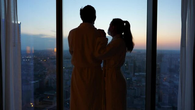 Young pensive couple waiting for quarantine or lockdown to end. Man and woman standing against the window in bath robes and facial masks. They look to each other then look out at evening city