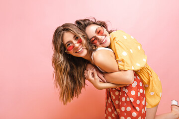 Young fair-skinned girl with closed eyes hugging her best friend from behind in pink studio. Blonde...