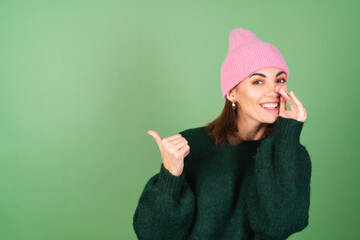 Young woman on a green background in a warm cozy sweater and a pink hat tells a secret, whispers,...